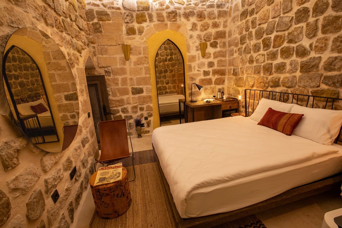 A stone-walled bedroom with a bed, a chair, a small table, and a mirror.