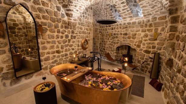 Un baño de paredes de piedra con una bañera independiente, un espejo y una pequeña mesa con velas y flores.
