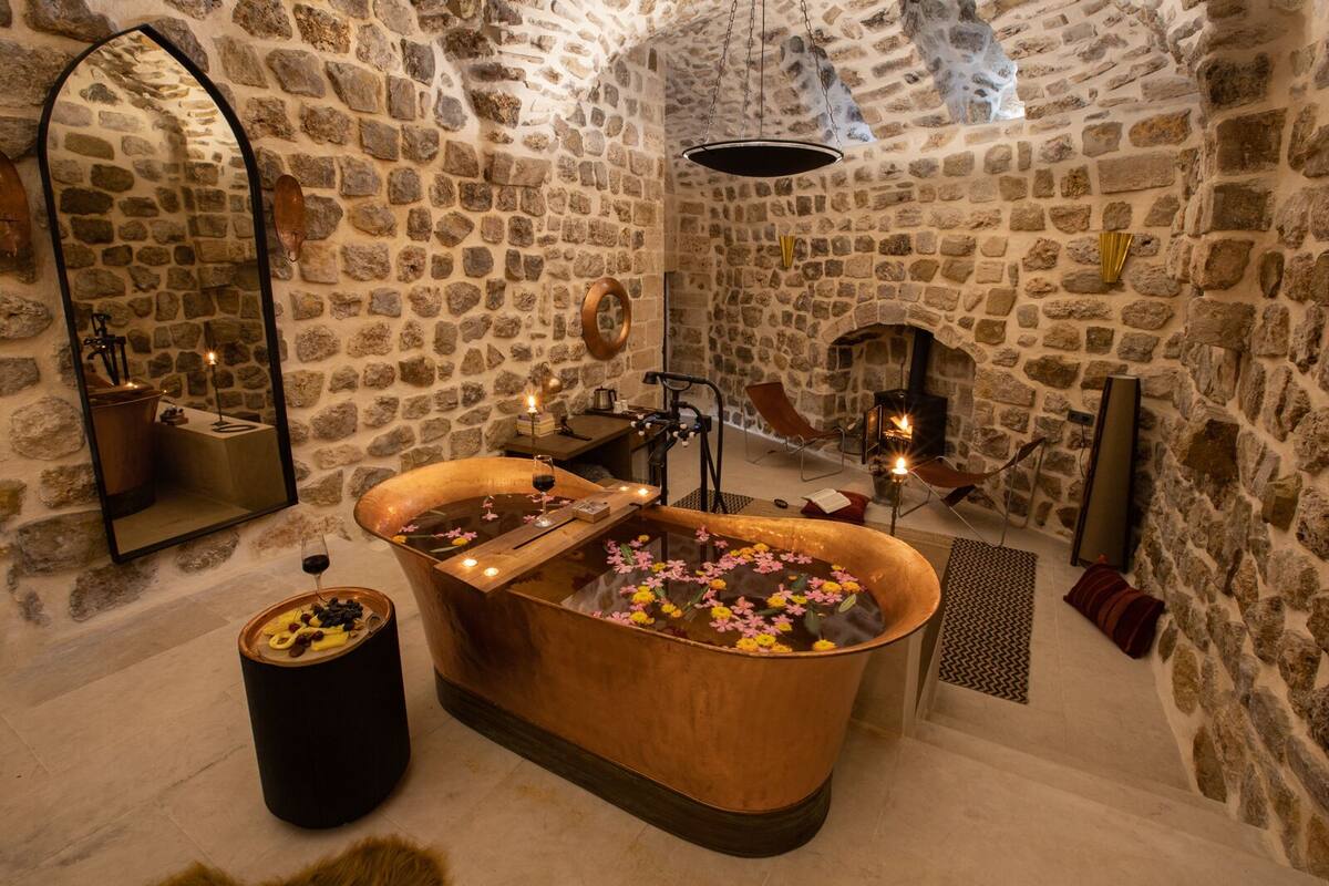 A stone-walled bathroom with a freestanding bathtub, a mirror, and a small table with candles and flowers.