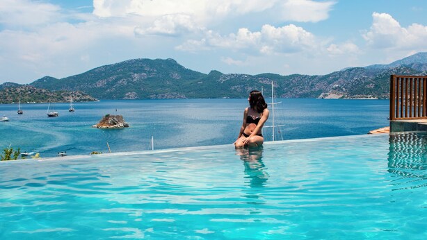 Una mujer sentada junto a una piscina azul cristalina, con vistas al mar y a las montañas.