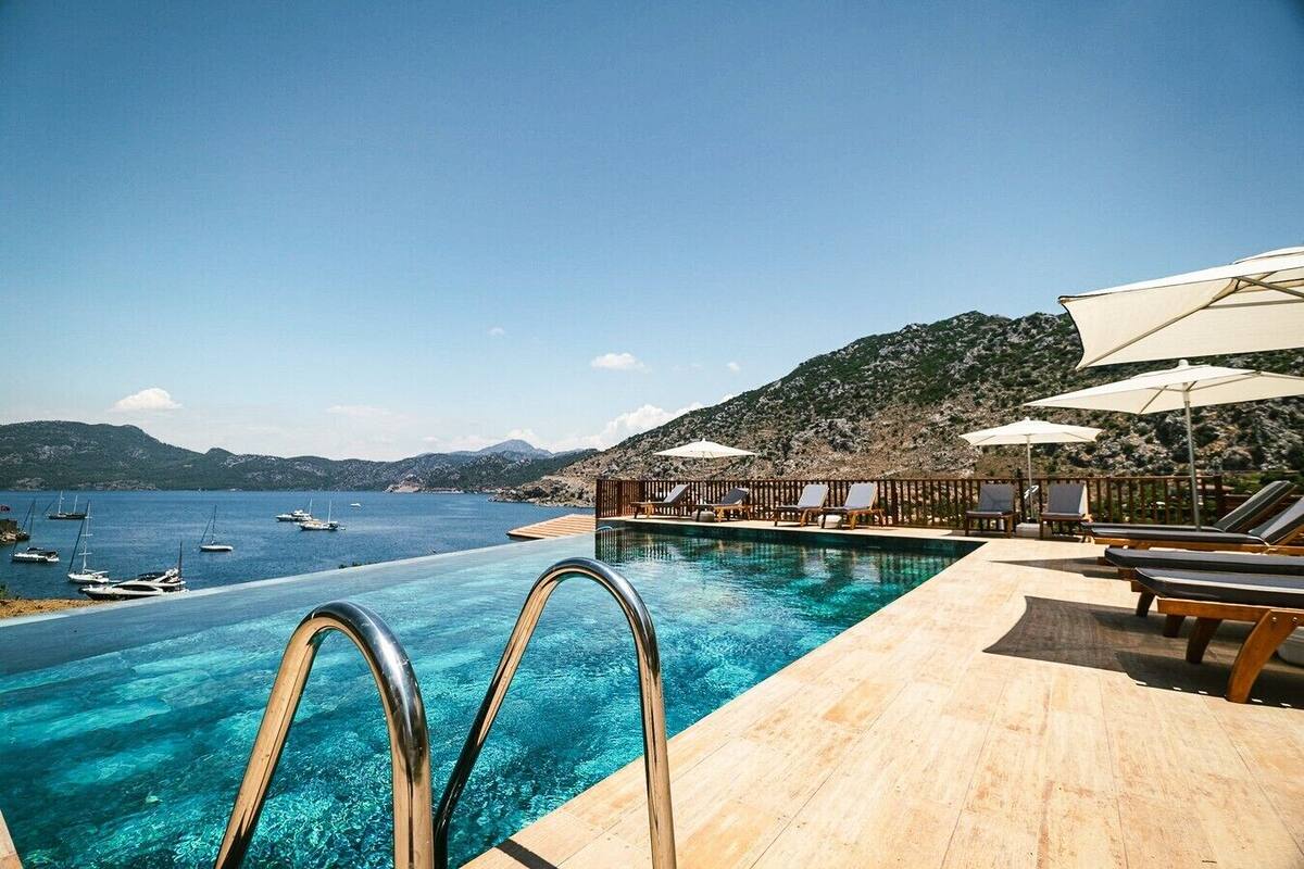 An infinity pool with clear blue water, lounge chairs, umbrellas, and a view of boats on the water.