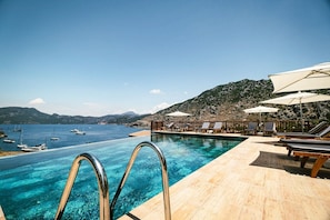 An infinity pool with clear blue water, lounge chairs, umbrellas, and a view of boats on the water.