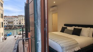 Ein Balkon mit Blick auf einen Kanal und Gebäude, ein Bett mit Kissen und Decke, und eine offene Tür, die zu einem Zimmer führt.