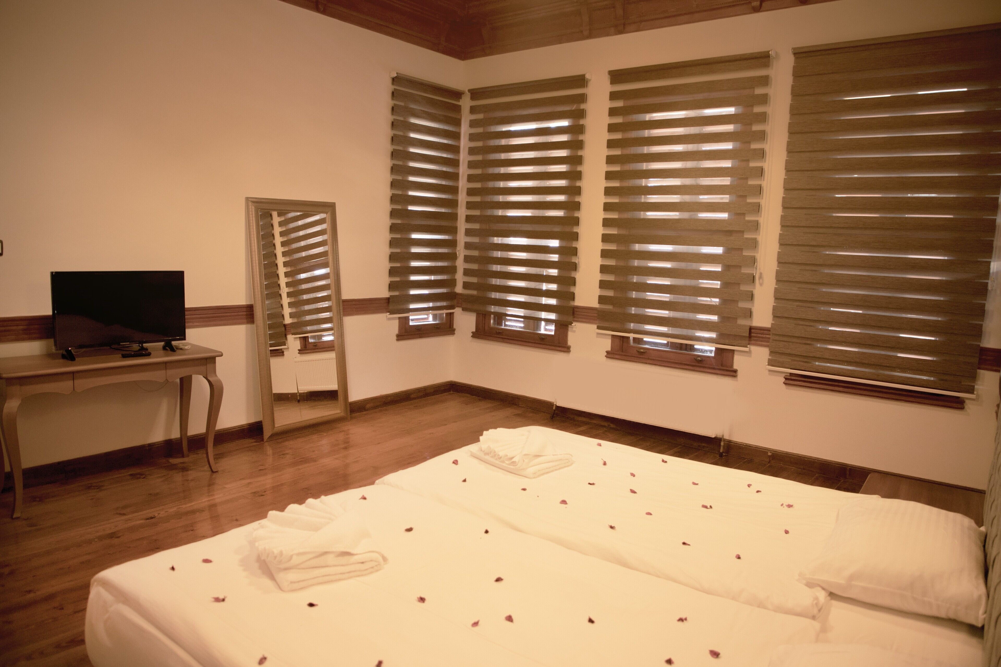 A hotel room with a large bed, a desk with a television, and three windows with blinds.