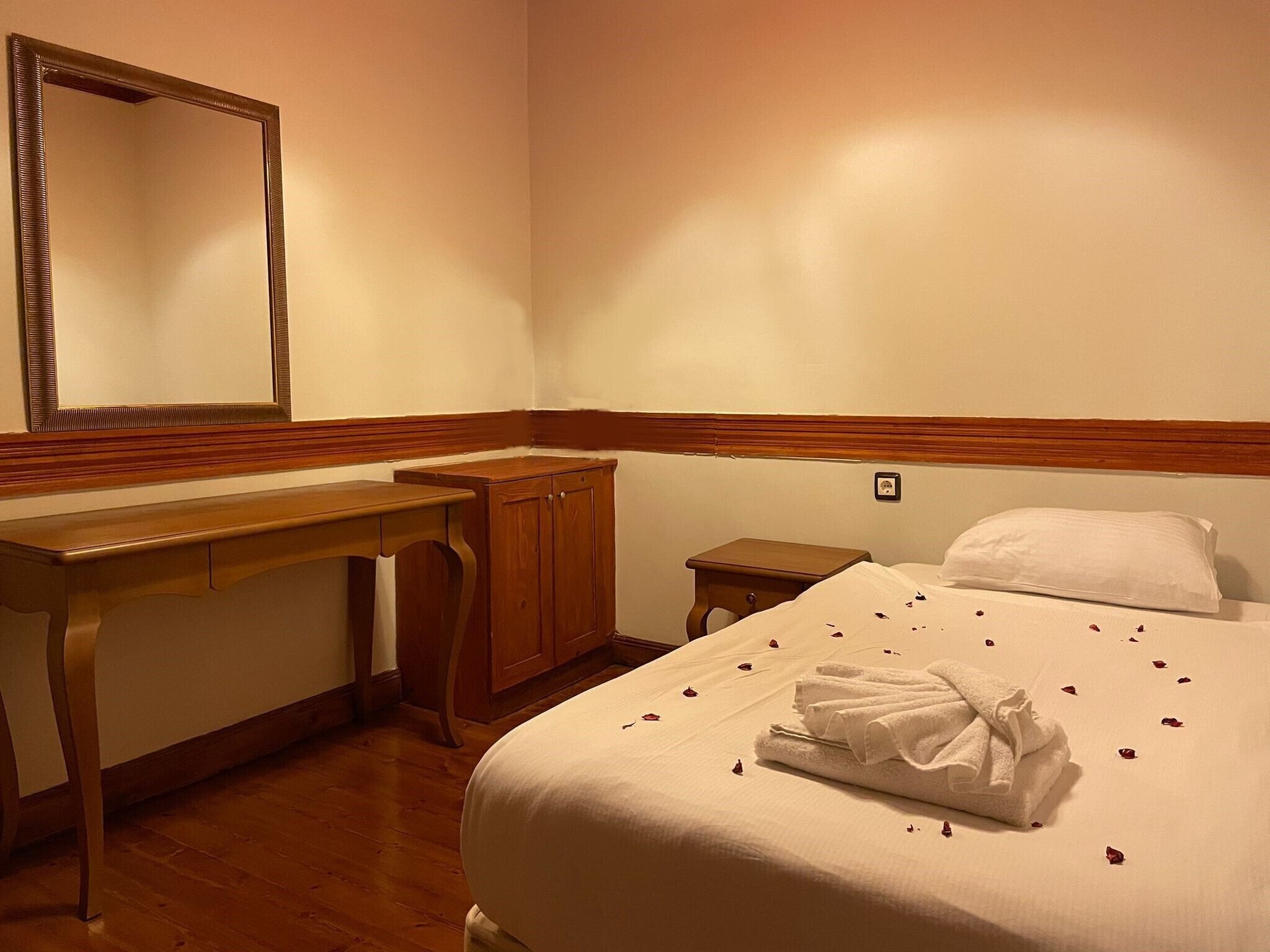 Habitació d'hotel amb escriptori de fusta, un armari, un llit amb roba blanca i pètals de rosa, i un mirall gran a la paret.