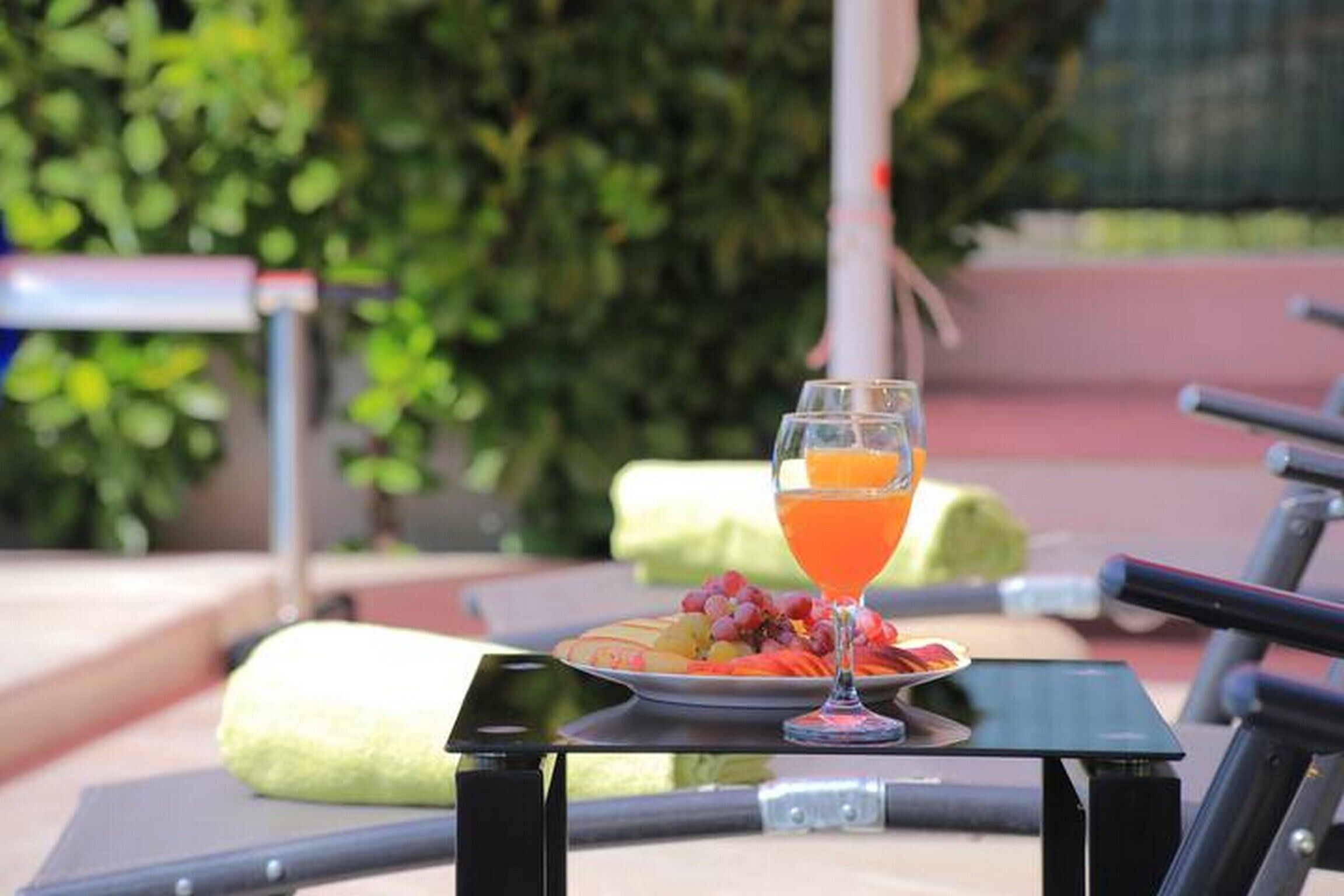 A table with a glass of orange juice and a plate of fruit, set on a sunny outdoor patio with chairs and greenery in the background.
