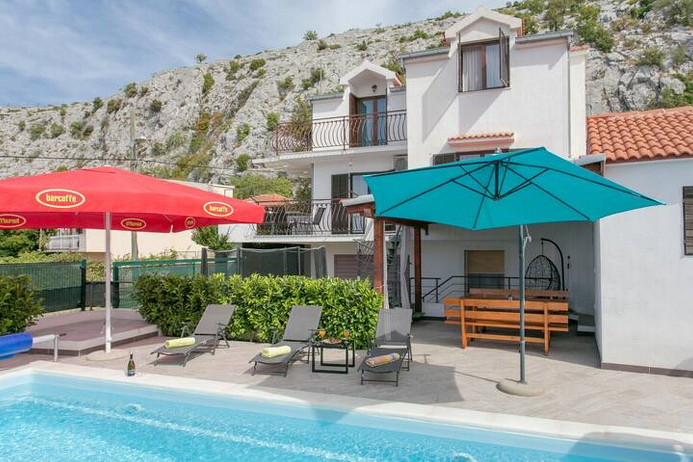 A pool area with lounge chairs and umbrellas, a white building with a balcony, and a rocky hillside in the background.