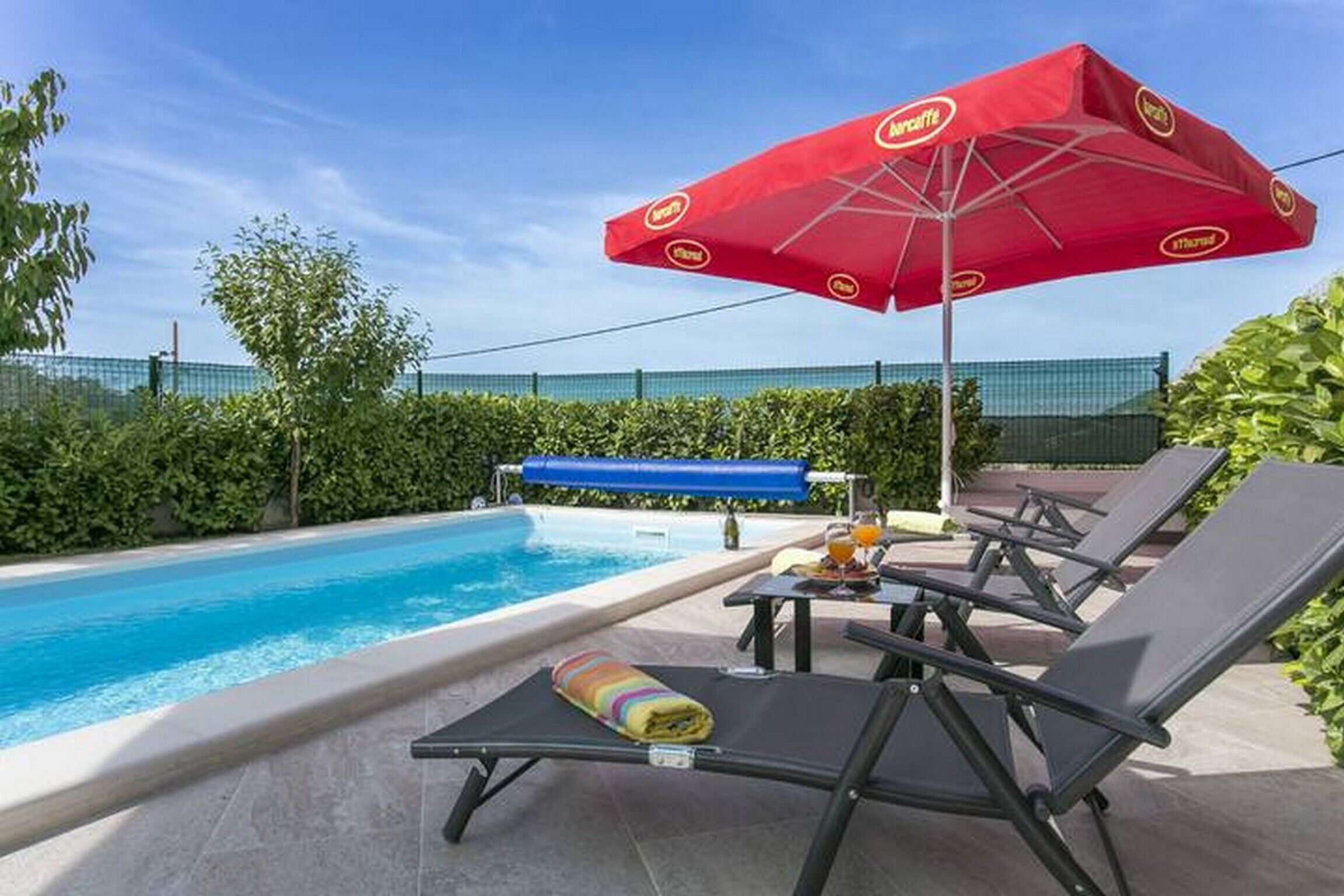 Zona junto a la piscina con sillones de descanso, una sombrilla roja y un toldo azul.