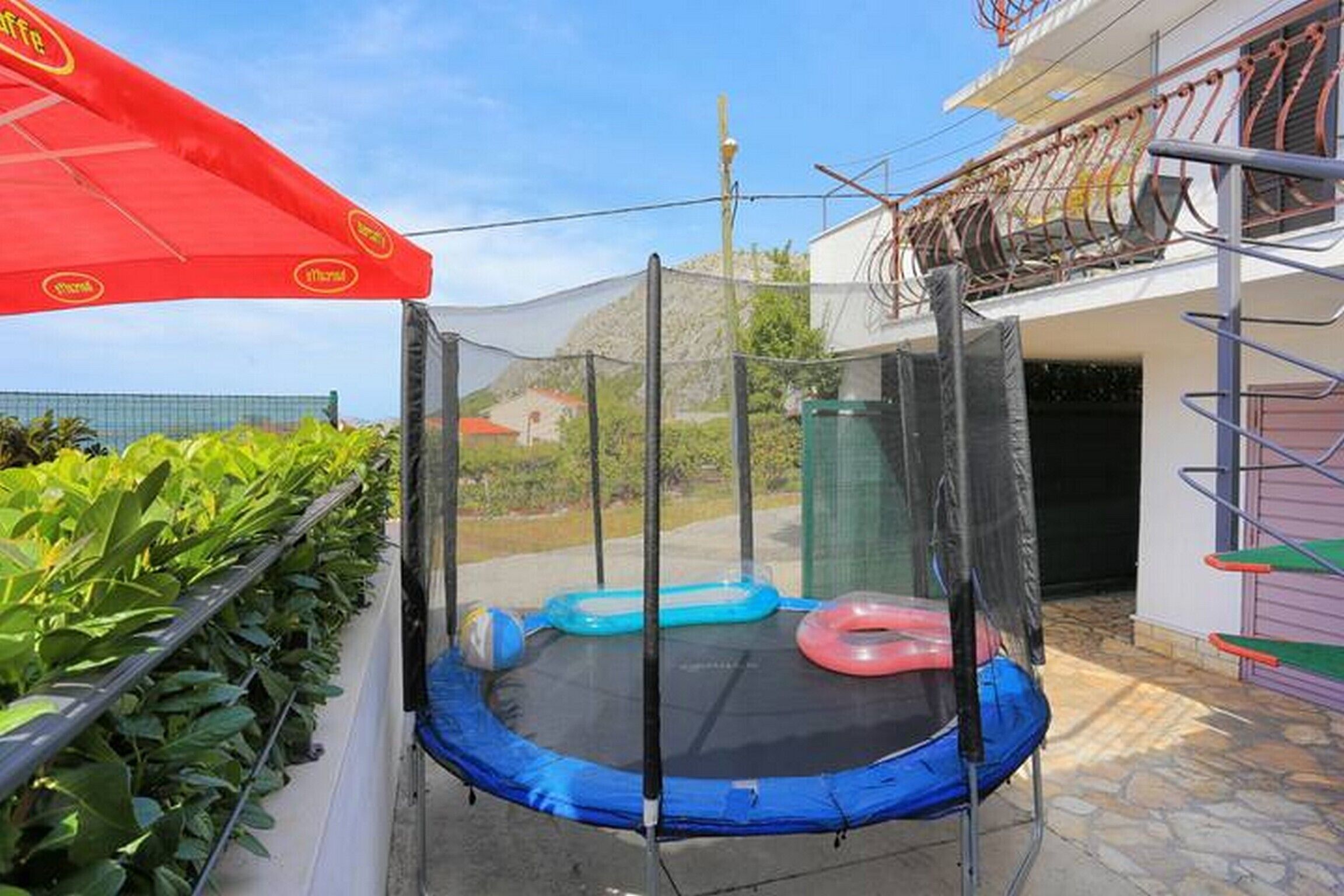A trampoline with safety netting, surrounded by a green hedge and a red umbrella.