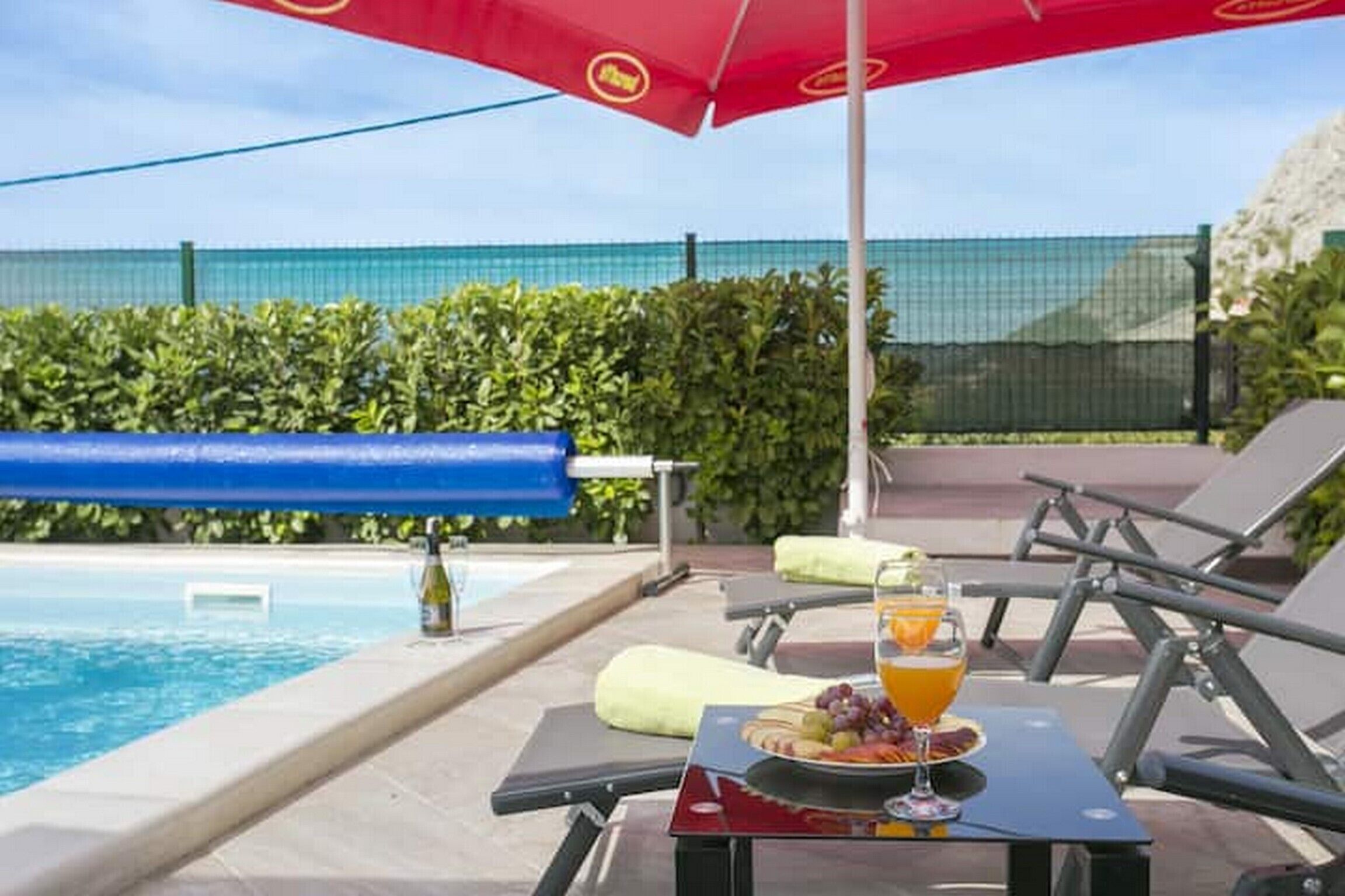 A poolside area with lounge chairs, a table with a drink and fruit, and a red umbrella.
