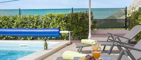A poolside area with lounge chairs, a table with a drink and fruit, and a red umbrella.