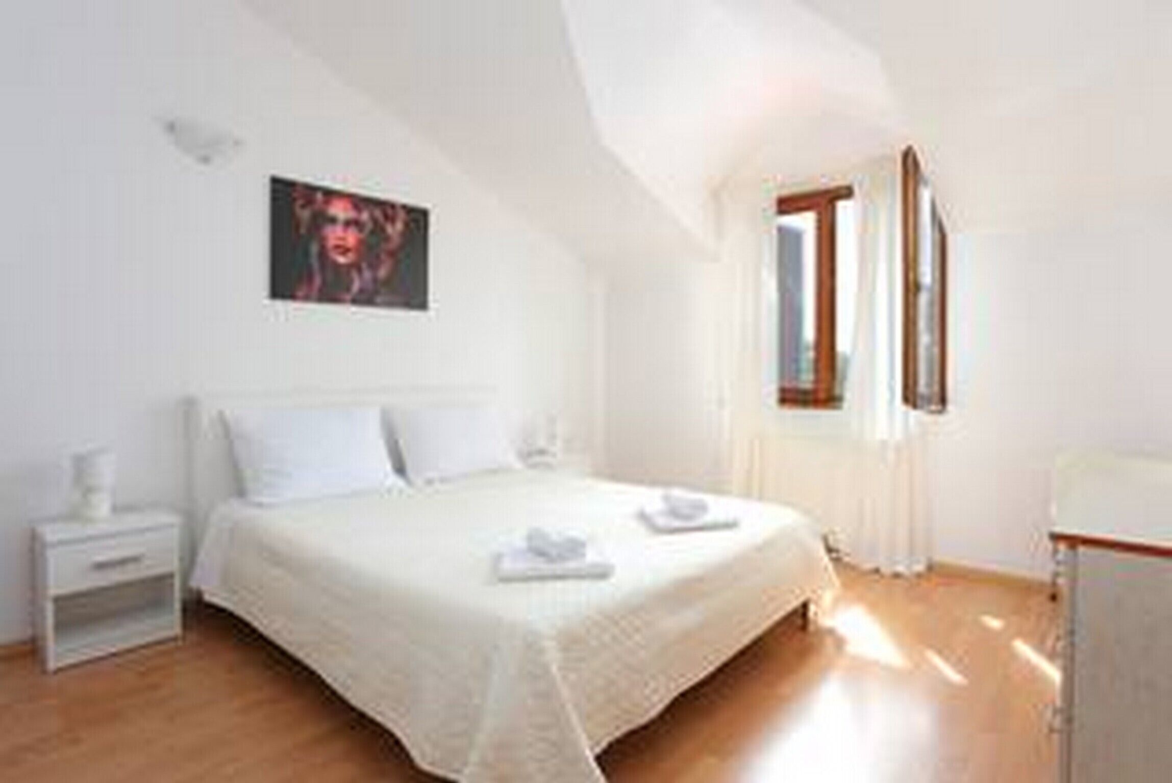 A bedroom with a bed, a nightstand, a window, and a painting on the wall.