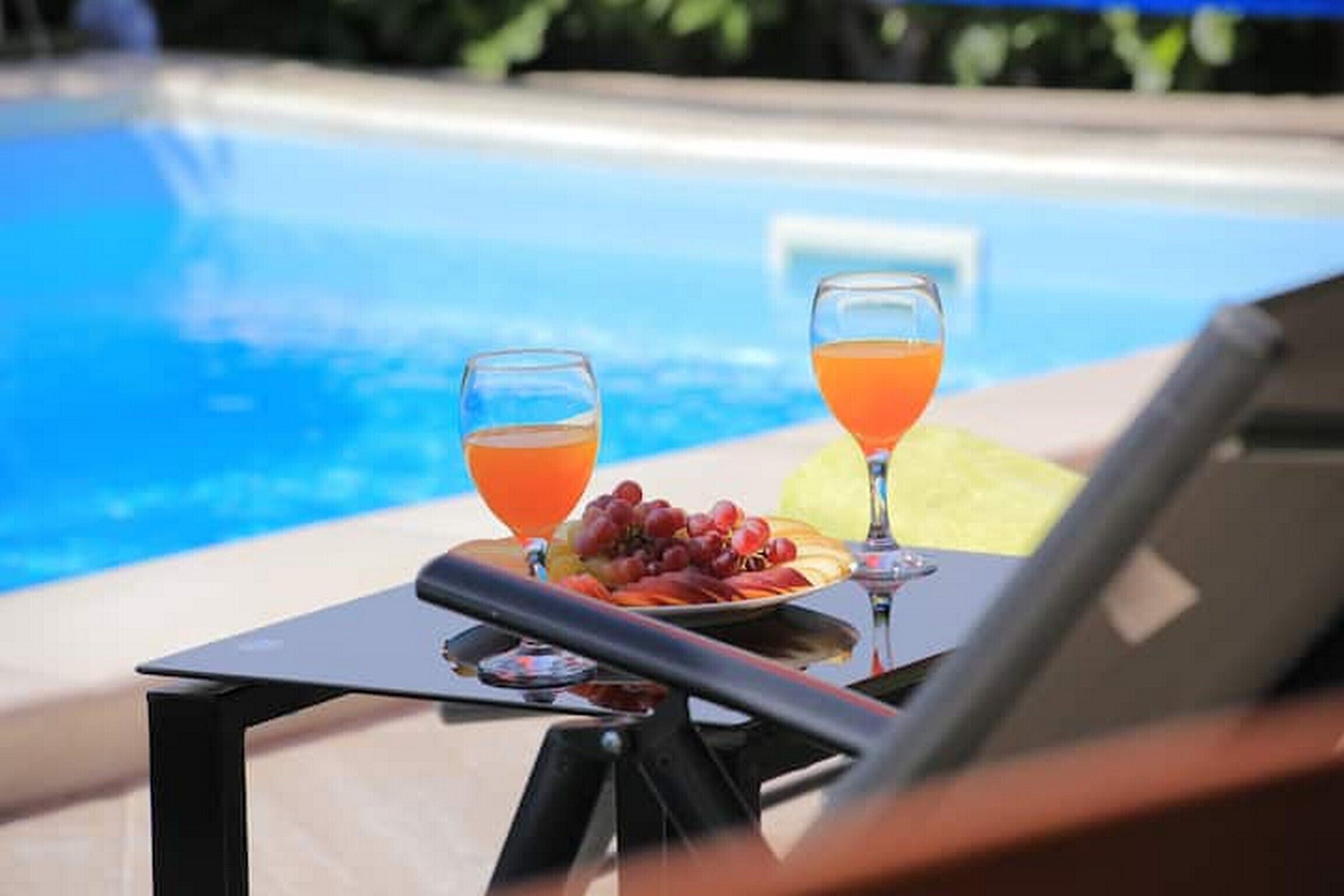 A poolside table with two glasses of orange juice and a plate of fruit.