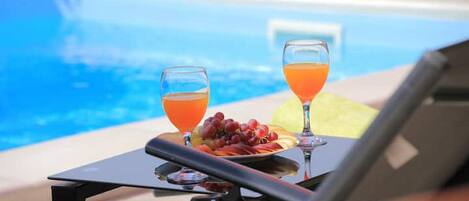 A poolside table with two glasses of orange juice and a plate of fruit.