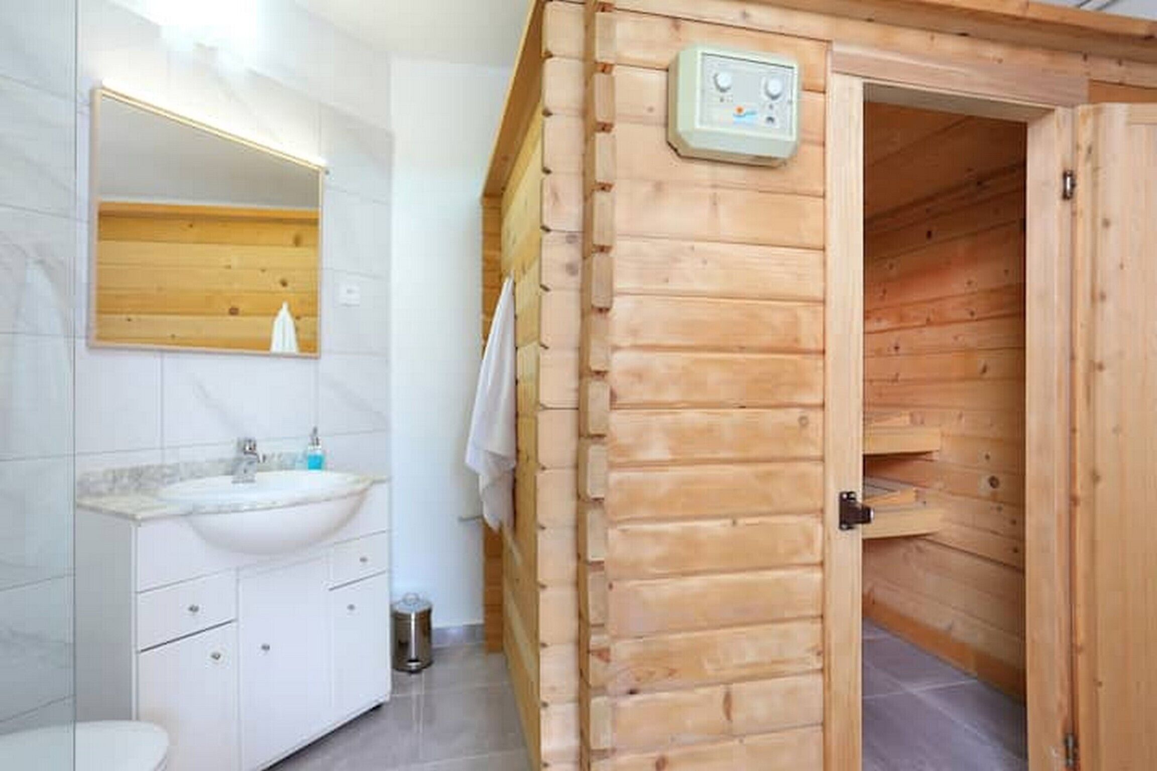 A wooden sauna with a control panel, a white sink, and a mirror in a modern bathroom.