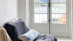 Ein Zimmer mit einem Bett, das mit einer dunklen Decke bezogen ist, einem Fenster mit Blick auf eine Landschaft und einer weißen Tür mit einem Metallgitter.