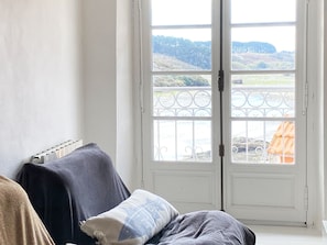 Ein Zimmer mit einem Bett, das mit einer dunklen Decke bezogen ist, einem Fenster mit Blick auf eine Landschaft und einer weißen Tür mit einem Metallgitter.