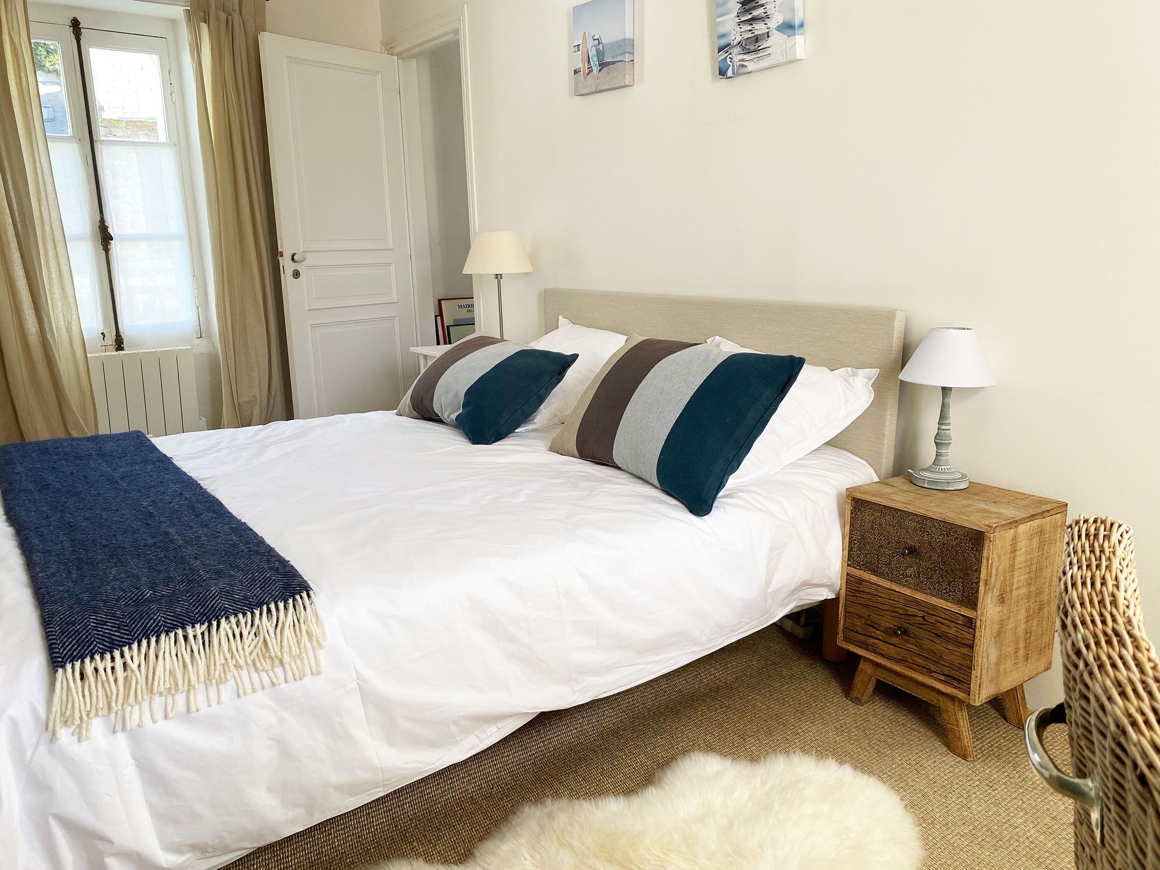 A neatly made bed with a white duvet and striped pillows, a wooden nightstand with a lamp, and a wicker chair.