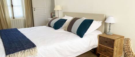 A neatly made bed with a white duvet and striped pillows, a wooden nightstand with a lamp, and a wicker chair.
