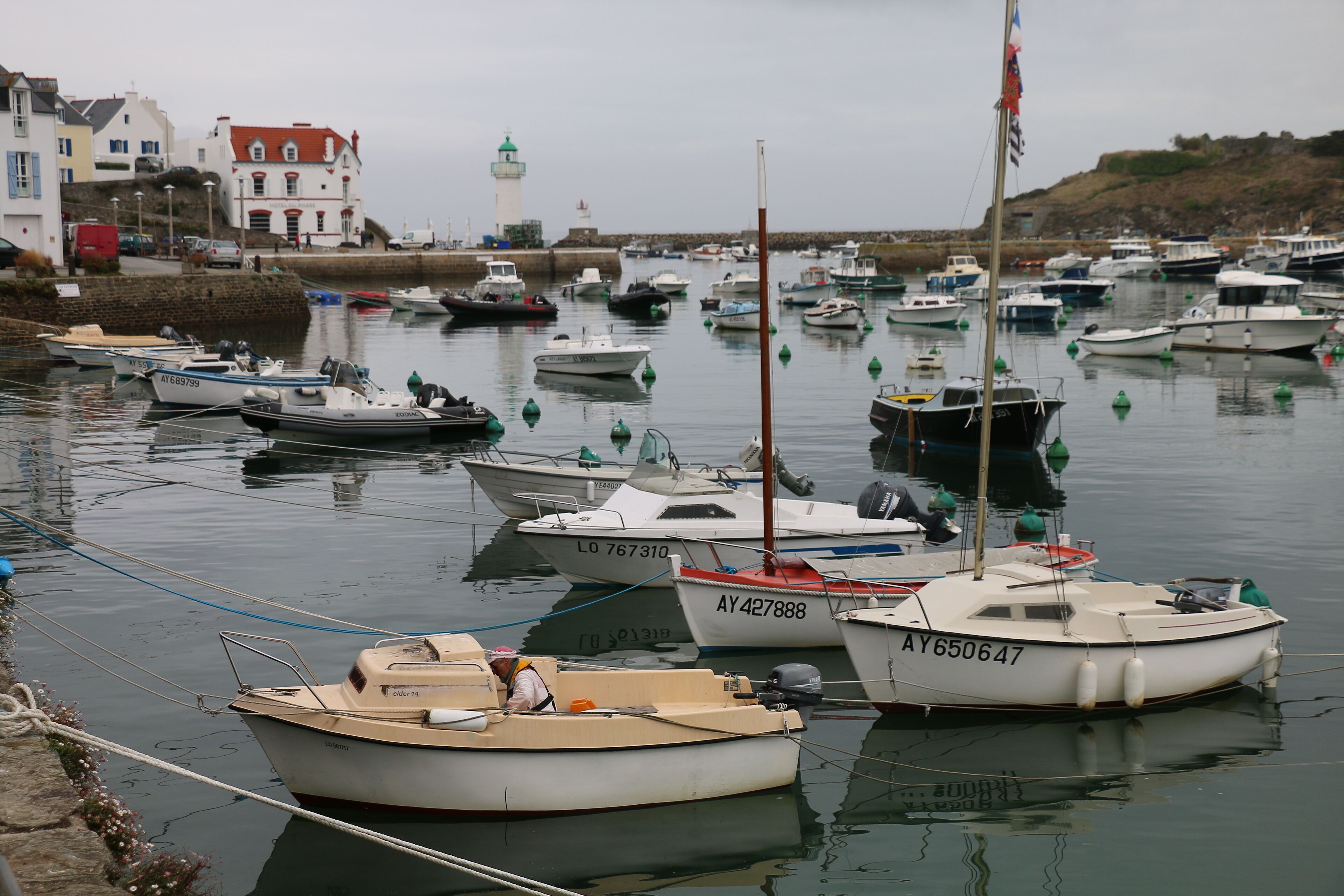 A marina with various boats docked, a lighthouse in the background, and a hotel nearby.