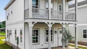 Une maison à deux étages avec un balcon à l’étage supérieur, un porche à l’avant et un petit jardin devant la maison, avec un arbre.