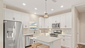 Une cuisine moderne avec des meubles blancs, des appareils en acier inoxydable, un îlot central doté de tabourets et un luminaire suspendu.