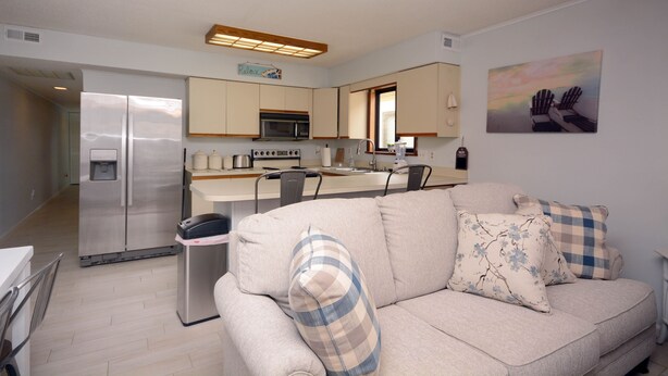 A modern kitchen with a refrigerator, microwave, and sink. There is a dining area with chairs and a table. A beige sofa with patterned cushions is in the living area. - 2-bedroom condo with an outdoor pool located uptown on the bayside in a peaceful, family-friendly neighborhood! (OCEAN CITY)