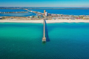Une vue aérienne d’une zone côtière, avec une longue jetée s’étendant dans la mer turquoise, entourée de bâtiments et d’une plage de sable.