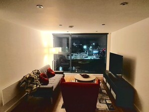 Ein Wohnzimmer mit einer Couch, einem roten Sessel, einem kleinen Tisch und einem Fernseher. Ein großes Fenster bietet einen Blick auf die beleuchtete Stadt bei Nacht.