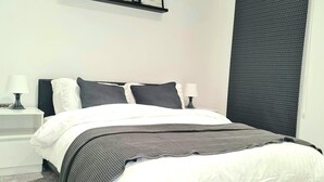 Een opgemaakt bed met wit en donkergrijs beddengoed, twee bedlampen en een aan de muur bevestigd fotolijstje.