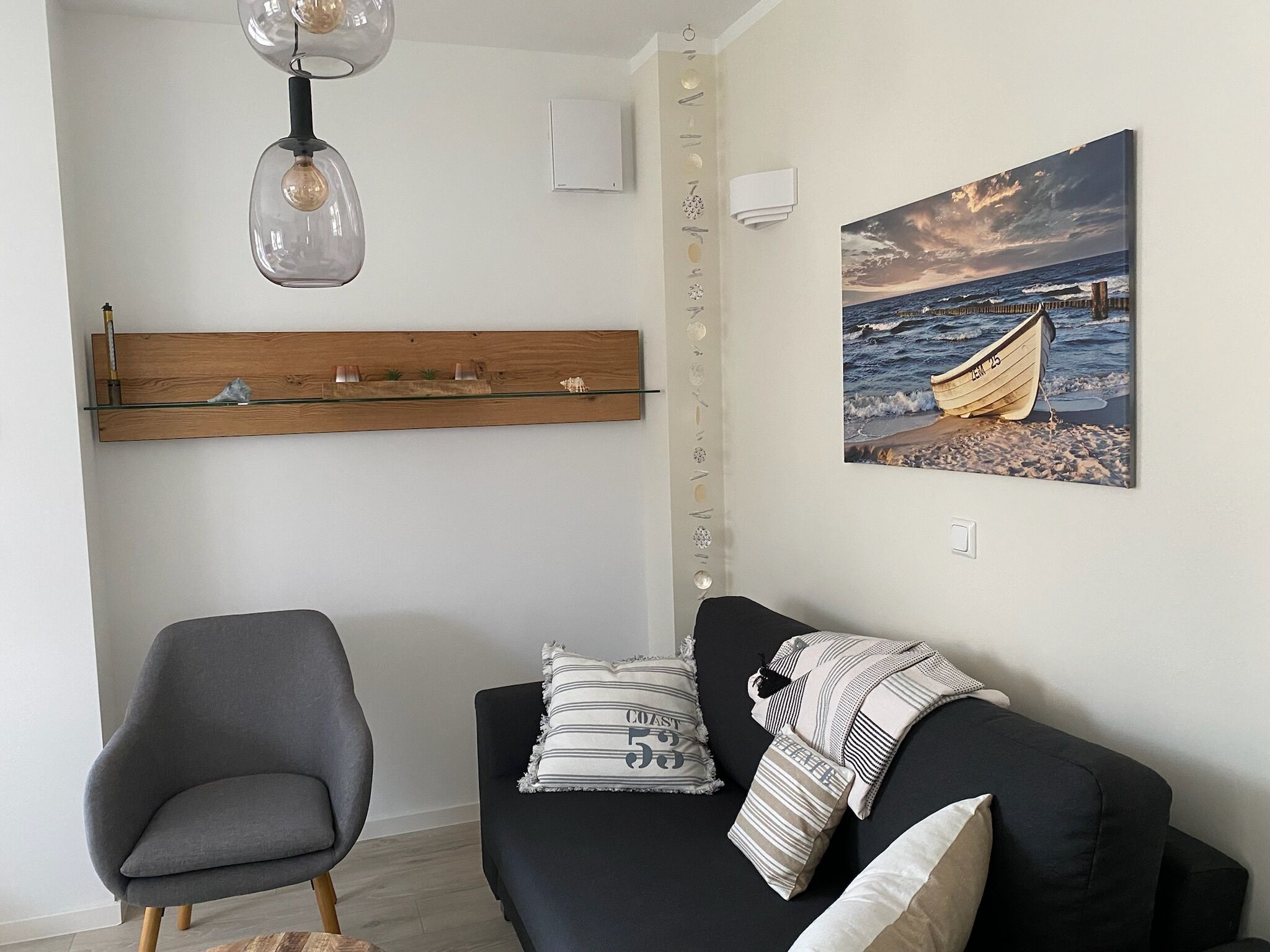 A living room with a black sofa, a grey armchair, a wooden shelf with decorative items, a painting of a boat on a beach, and a pendant light.