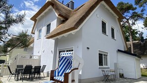 A two-story house with a thatched roof, a covered outdoor seating area with striped cushions, and a small shed. - Vacation home under thatch on Usedom for 6 people with sauna and wood-burning stove, wall box. (Zirchow)