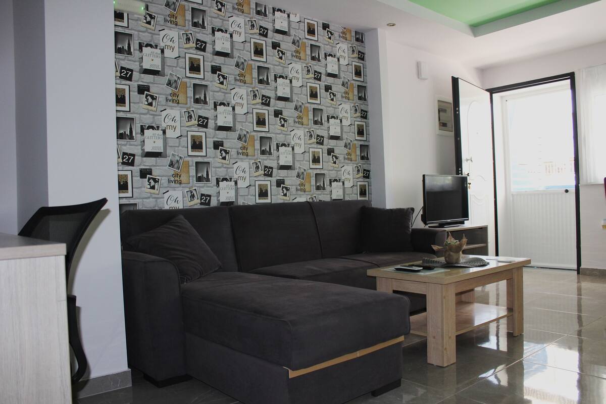 Ein modernes Wohnzimmer mit einer dunkelgrauen Eckcouch, einem Holztisch und einem Fernseher. Die Wand ist mit strukturierter Tapete verziert, die Schwarz-Weiß-Fotos und Text zeigt.