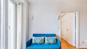 A blue sofa with floral cushions in a room with wooden flooring, white walls, and a door leading to another room. - 2BR at Chiado Historical Heart of Lisbon (Lisbon)