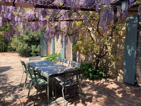 An outdoor patio with a table and chairs, surrounded by a stone wall and covered with purple wisteria.