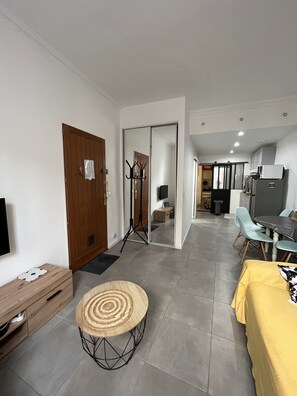 Un salon moderne avec une porte en bois, un téléviseur, une petite table ronde dont le plateau est fait d’un matériau tressé, et un coin repas avec une nappe jaune.
