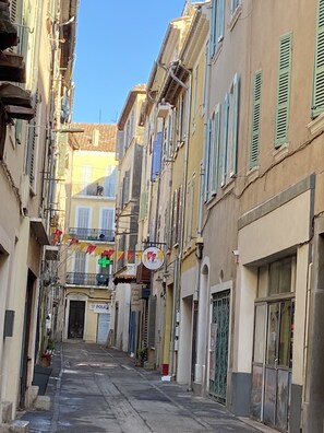Une rue étroite bordée d’immeubles à plusieurs étages, dont certains sont dotés de balcons et de volets.