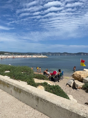 A beach with people sunbathing and swimming, a flag, and boats in the distance.