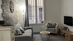 A modern living room with a brick wall, a sofa, a round coffee table, a TV, and a window with a view of buildings. - TYPE2, a rare 4-person gem, ideally renovated (La Ciotat)