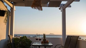Un balcón con una mesa dispuesta para dos personas, que incluye dos copas de vino tinto, una piña y un frutero. Se observa el océano y una montaña distante.