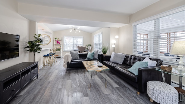 A spacious living room with a dark leather sofa, a coffee table, and a TV on a wooden cabinet. There are two windows with blinds, a chandelier, and a potted plant. - 1 Block to Allianz Field • Sleeps 8+ • Patio & Parking (Saint Paul)