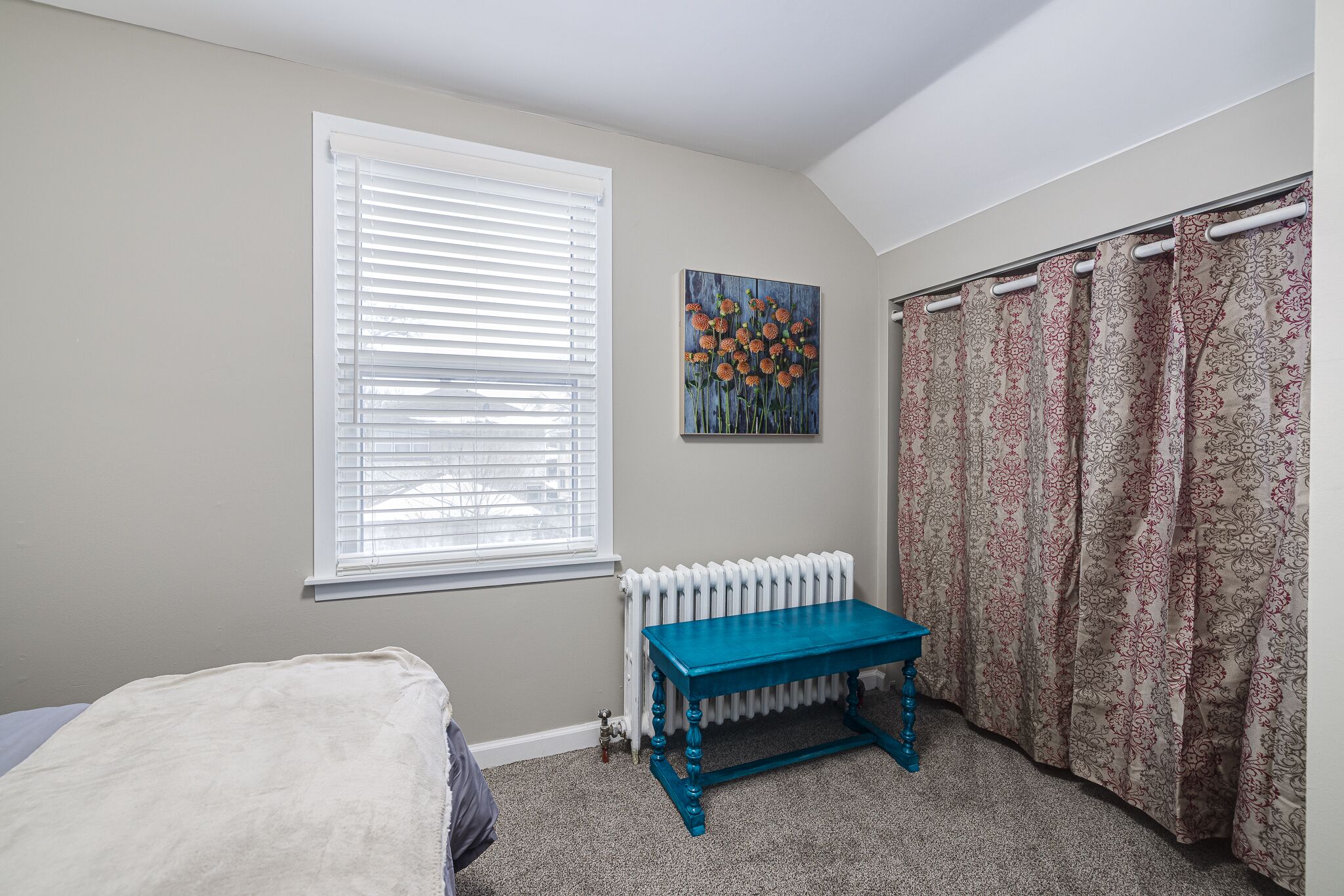 A bedroom with a bed, a blue table, a radiator, a window with blinds, a painting, and patterned curtains.
