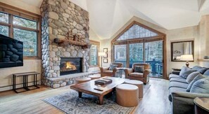 Amplia sala de estar con chimenea de piedra, asientos confortables y ventanas grandes con vistas al exterior.
