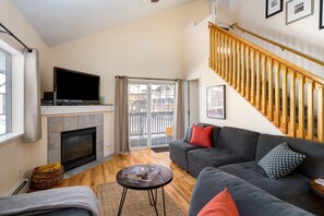 A modern living room with a fireplace, a large TV, a grey sofa, and a wooden staircase. - Quail Run 5 3 Bedroom Condo by RedAwning (Steamboat Springs)