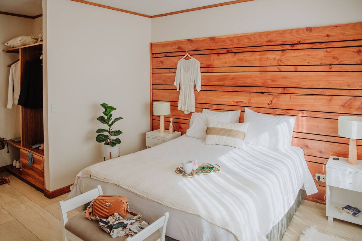 Une chambre à coucher avec une tête de lit en bois, un lit avec une literie blanche, une table de chevet avec une lampe, un peignoir suspendu et une plante en pot.