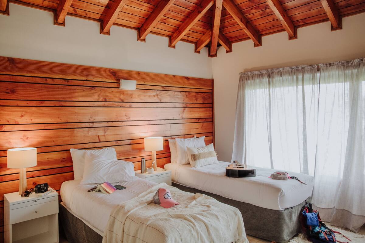 Une chambre avec deux lits, des poutres apparentes en bois au plafond, une table de chevet avec une lampe et une fenêtre ornée de rideaux légers.
