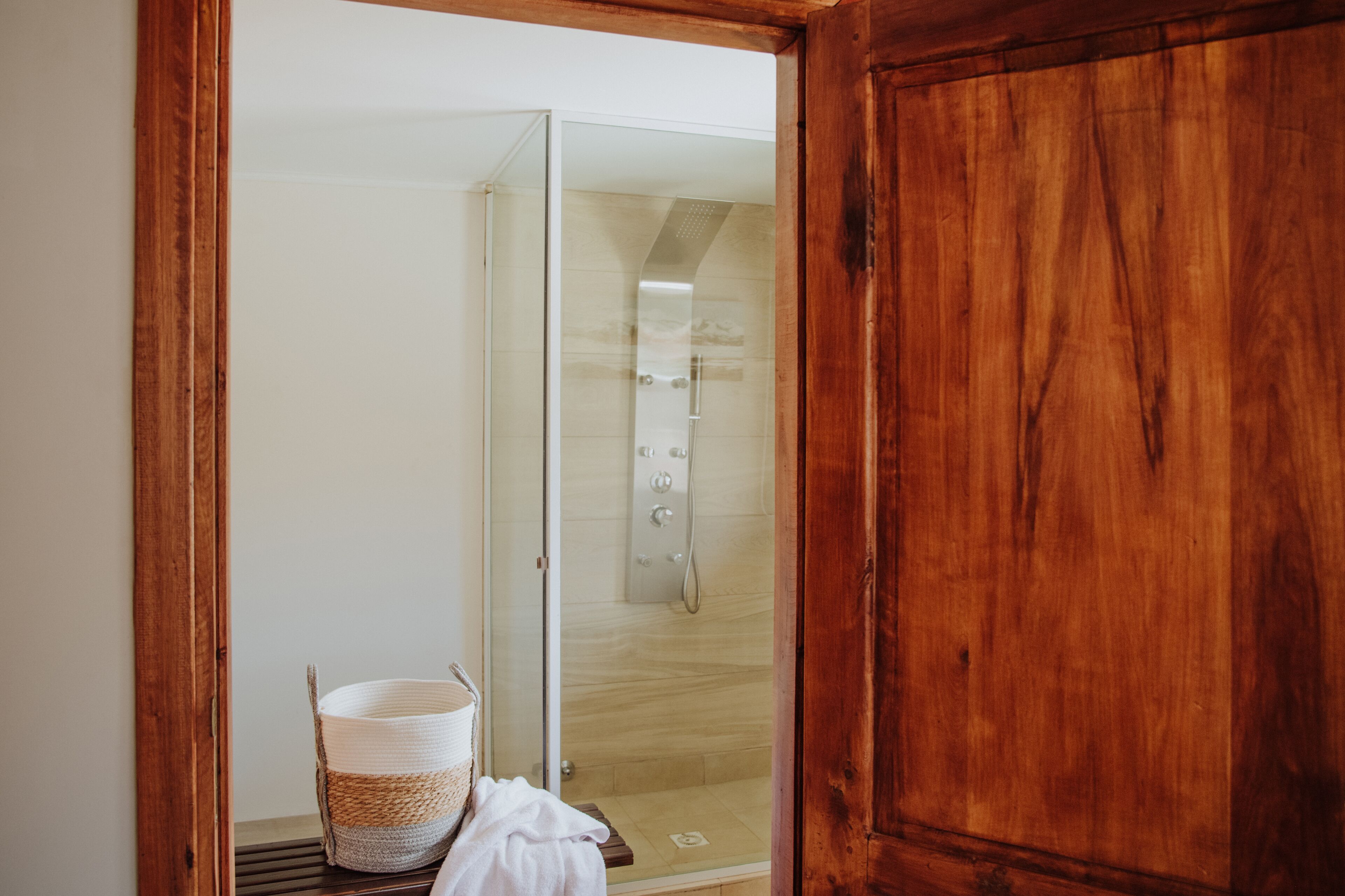 A wooden door leading to a glass shower area with a white towel and a woven basket on a wooden shelf.