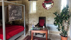 Una habitación rústica con techo de madera, una cama con colcha roja, una silla, una mesa con piezas de ajedrez, un espejo con forma de corazón y una planta en maceta.