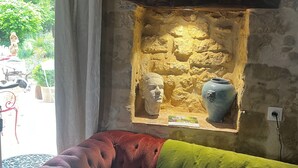 Una habitación con pared de piedra, una ventana con cortinas, un sofá rojo, un cojín verde y una estantería con objetos decorativos.