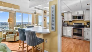 A modern kitchen with white cabinets, a stainless steel oven, and a microwave. There is a bar area with two striped barstools and a large window offering a city view. - Capri 7: 2 BR, 2 BA Condominium in Ocean City, Sleeps 6 (Ocean City)